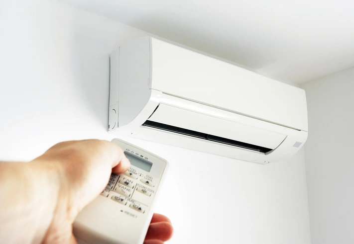 A person holding a remote control while standing next to an air conditioner, adjusting the temperature settings